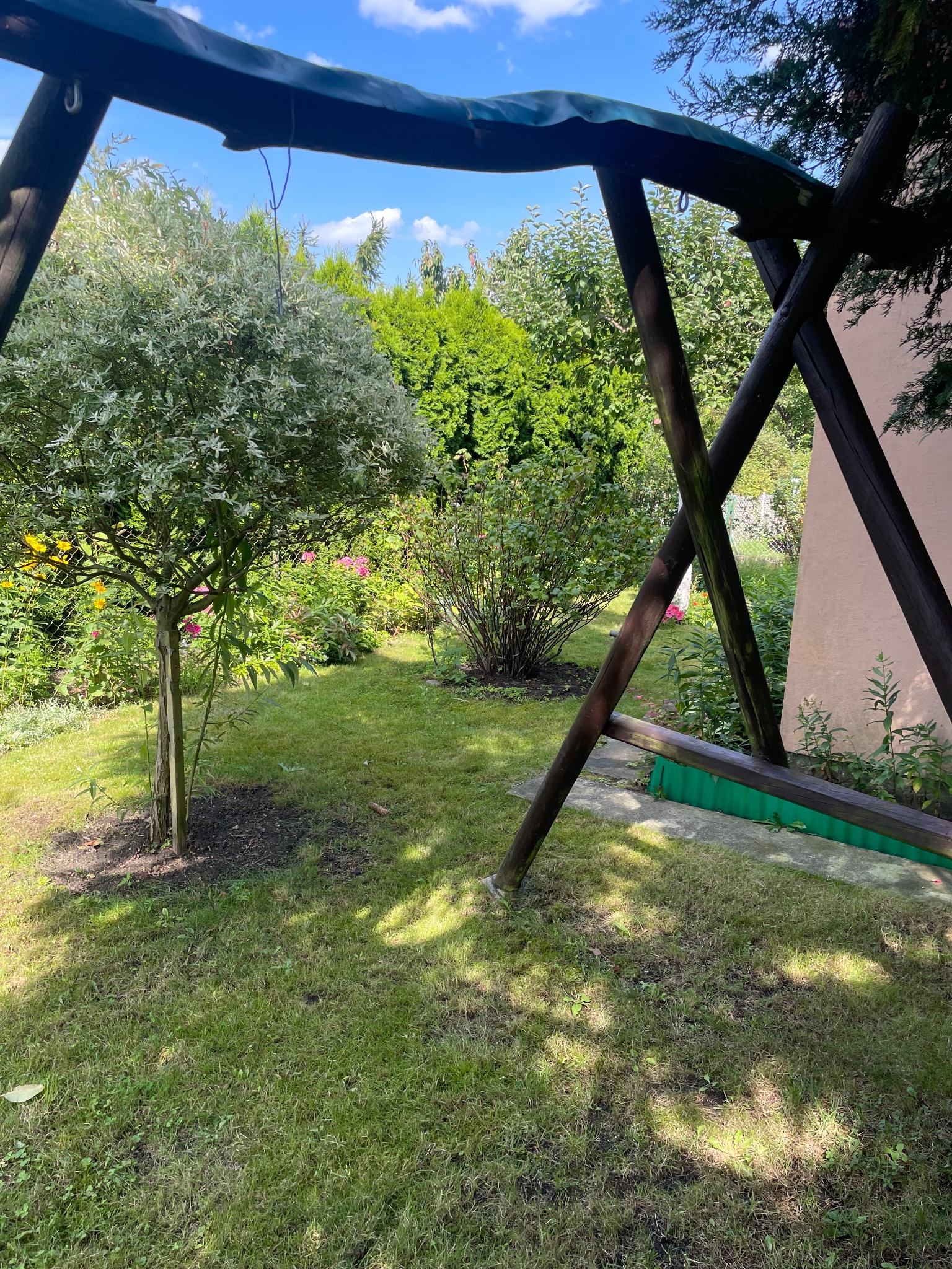 Zadbany ogród z zielonym trawnikiem, drzewami i huśtawką w słoneczny dzień. Widoczne kwiaty i krzewy. Elementy drewniane huśtawki w pierwszym planie.