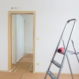Mariusz Zieliński - Białe ściany po malowaniu, zabezpieczone futryny, drabina z wałkiem i pędzlem. Podłoga zabezpieczona kartonem. Widok na sąsiednie pomieszczenie.
