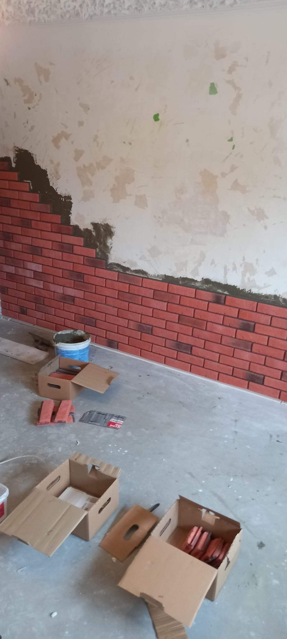 Montaż dekoracyjnej cegły na ścianie w trakcie prac. Widoczne pudełka z cegłami, wiadro z zaprawą i narzędzia. Częściowo wykończona ściana z cegły.