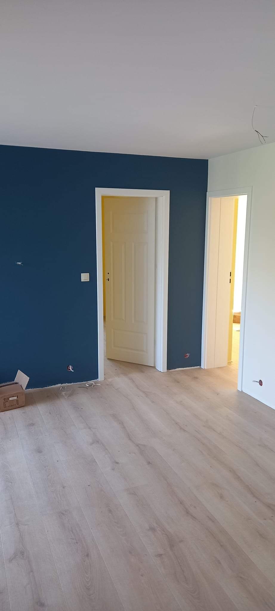 Wnętrze po remoncie: granatowa ściana, jasne panele podłogowe, białe drzwi. Widoczne gniazdka elektryczne i karton na podłodze. Dwa otwarte przejścia do innych pomieszczeń.
