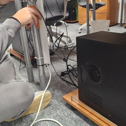 ALISON Krzysztof Banasik - Instalator mocuje kable pod biurkiem w biurze, używając opasek zaciskowych. Widoczny komputer stacjonarny, kable, narzędzia i fragment podłogi biurowej.