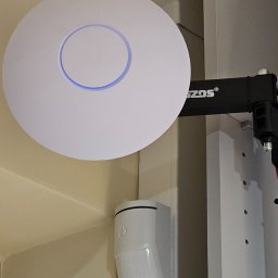 ALISON Krzysztof Banasik - Zamontowany punkt dostępowy Wi-Fi (access point) z niebieskim podświetleniem, obok czujnik ruchu. Widoczne elementy montażowe.