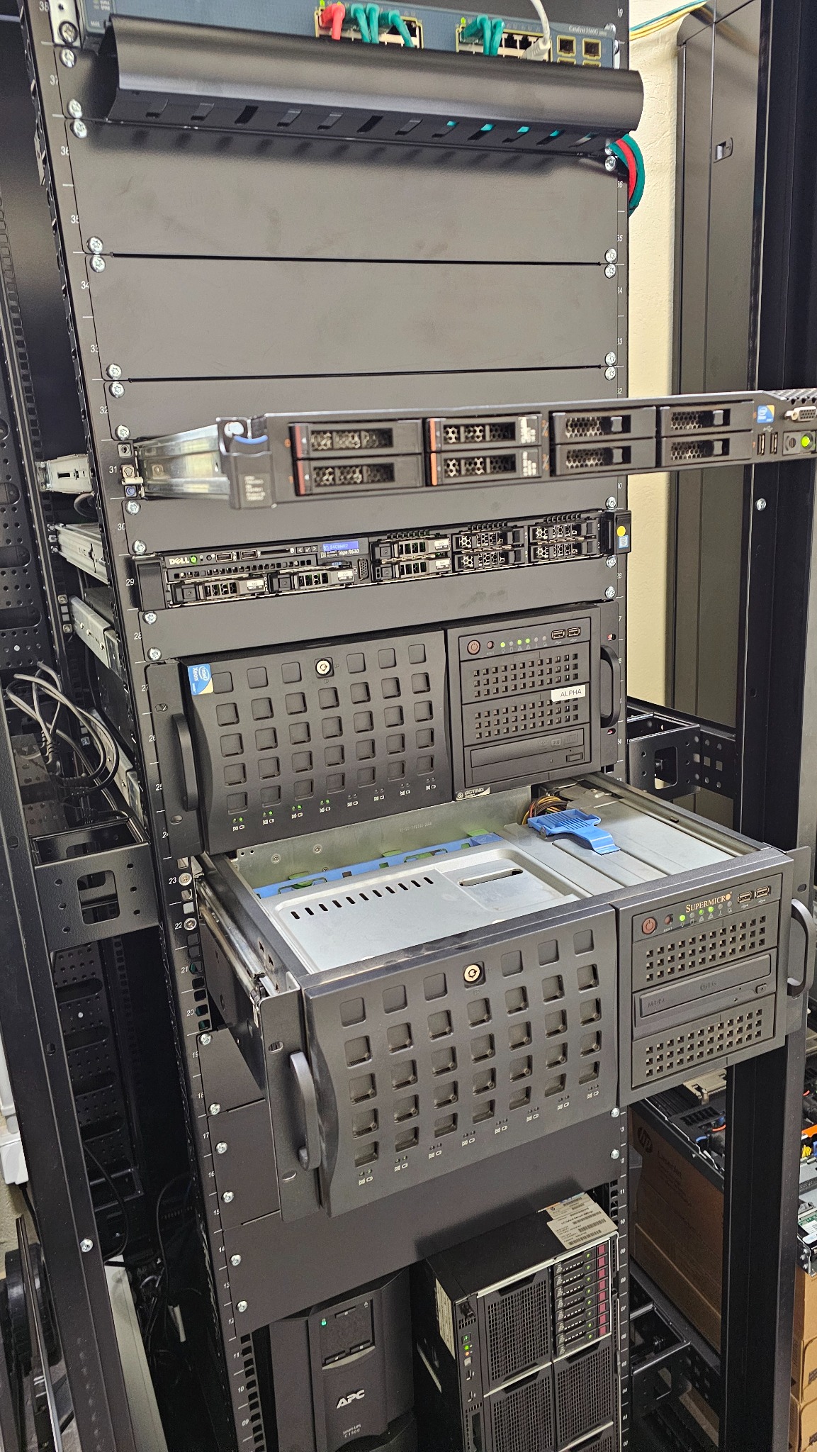 Serwerownia z szafą rack, w której zamontowane są serwery różnych producentów. Widoczne kable i zasilacze awaryjne APC. Jeden z serwerów jest wysunięty.