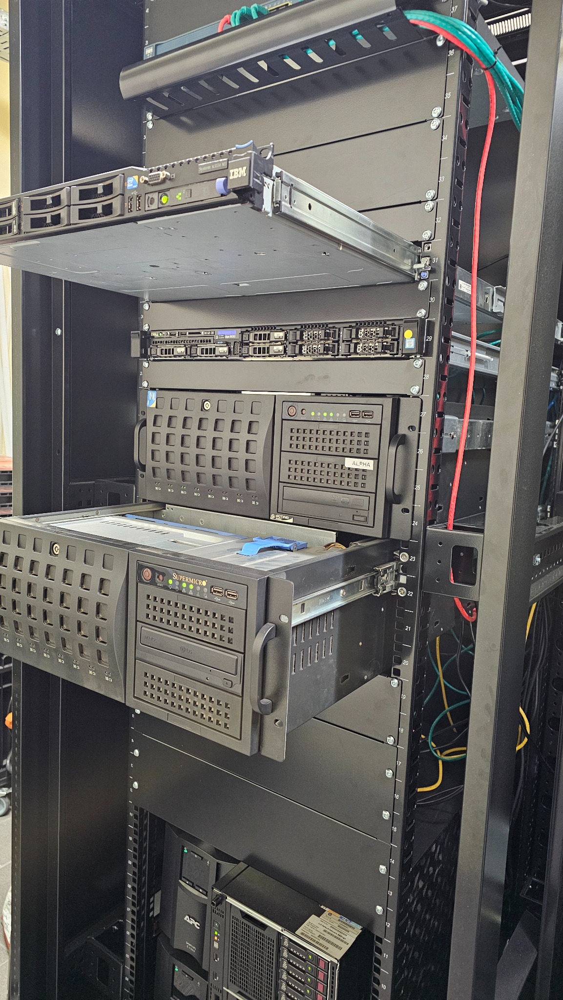 Serwerownia z widocznymi serwerami IBM i Supermicro w szafie rack. Kable sieciowe w górnej części. Widoczne numery na szafie rack.