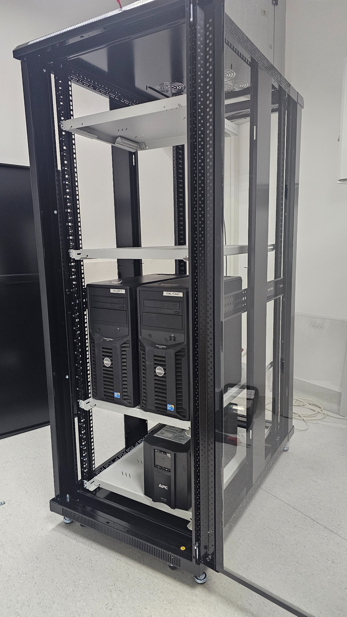 Serwerownia: szafa rack z dwoma komputerami Dell z naklejkami 'Hurt' i 'Detal' oraz zasilaczem awaryjnym APC. Widok z boku, oświetlenie studyjne.