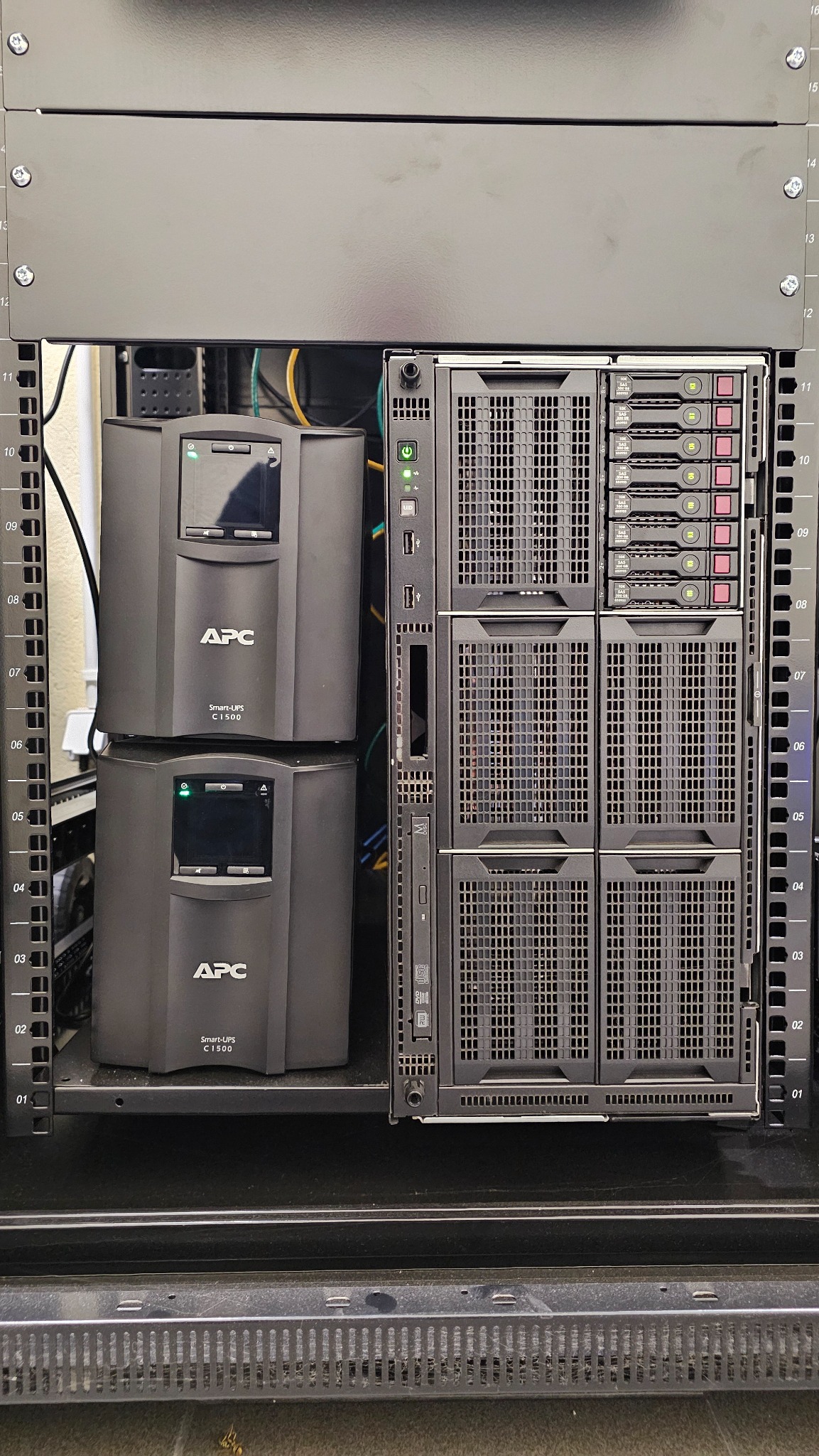 Serwerownia: Dwa zasilacze awaryjne APC Smart-UPS C1500 obok serwera w szafie rack. Widoczne kable i diody LED. Sprzęt IT w ciemnej obudowie.