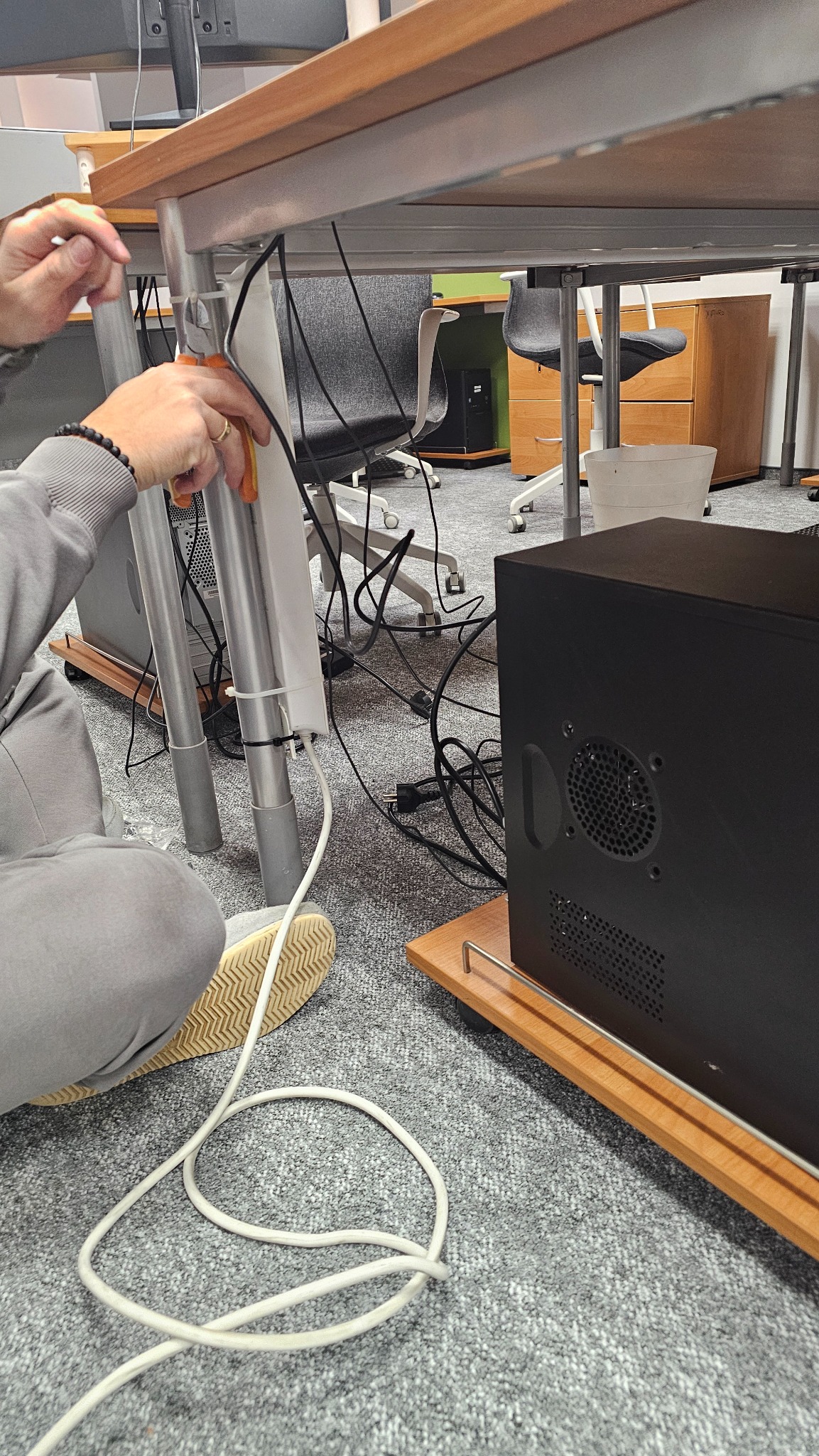 Instalator mocuje kable pod biurkiem w biurze, używając opasek zaciskowych. Widoczny komputer stacjonarny, kable, narzędzia i fragment podłogi biurowej.
