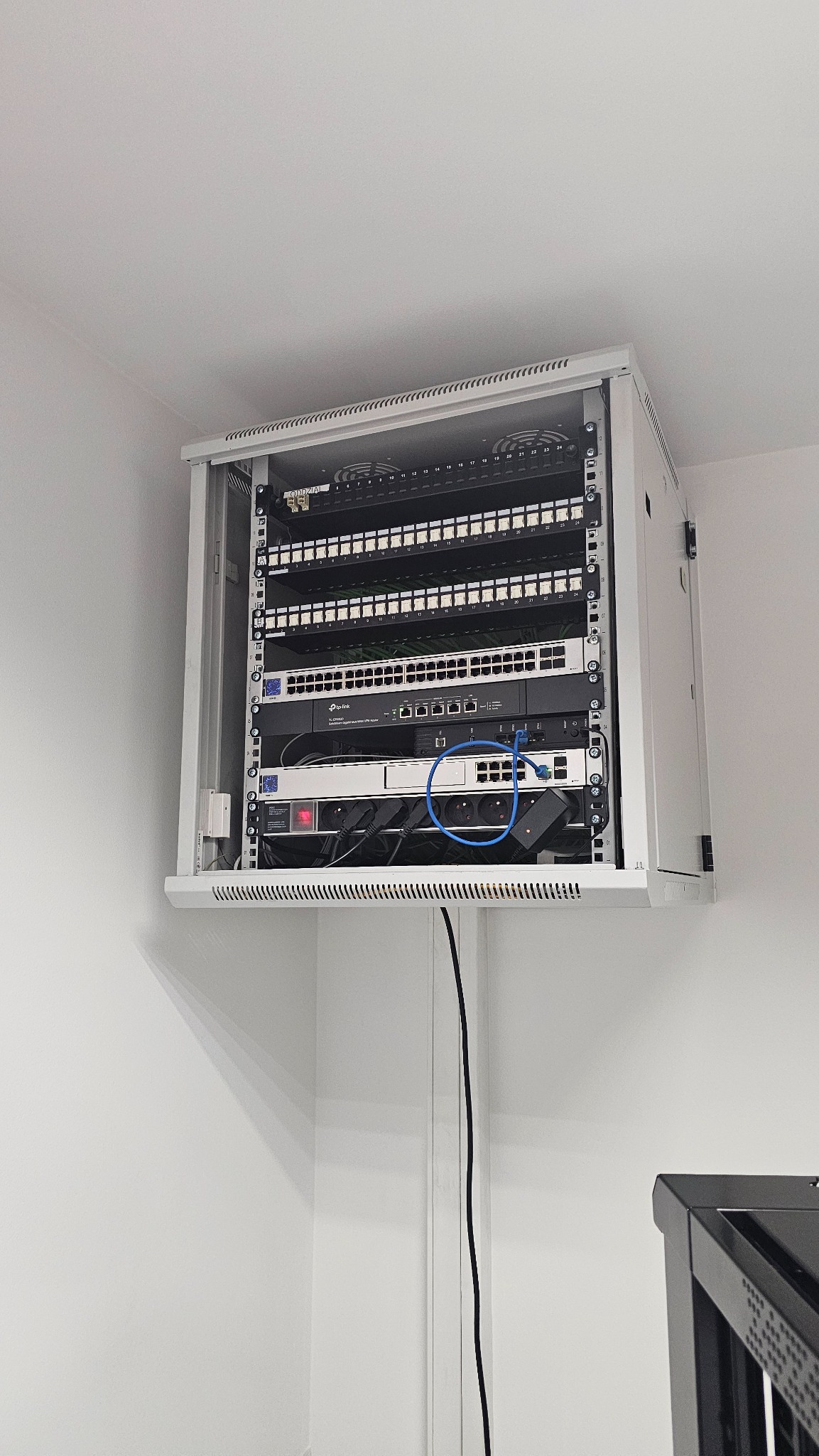 Zamknięta szafa rack z patch panelem, switchem i listwą zasilającą. Widoczne kable połączeniowe. Szafa zamontowana na ścianie w pomieszczeniu biurowym.