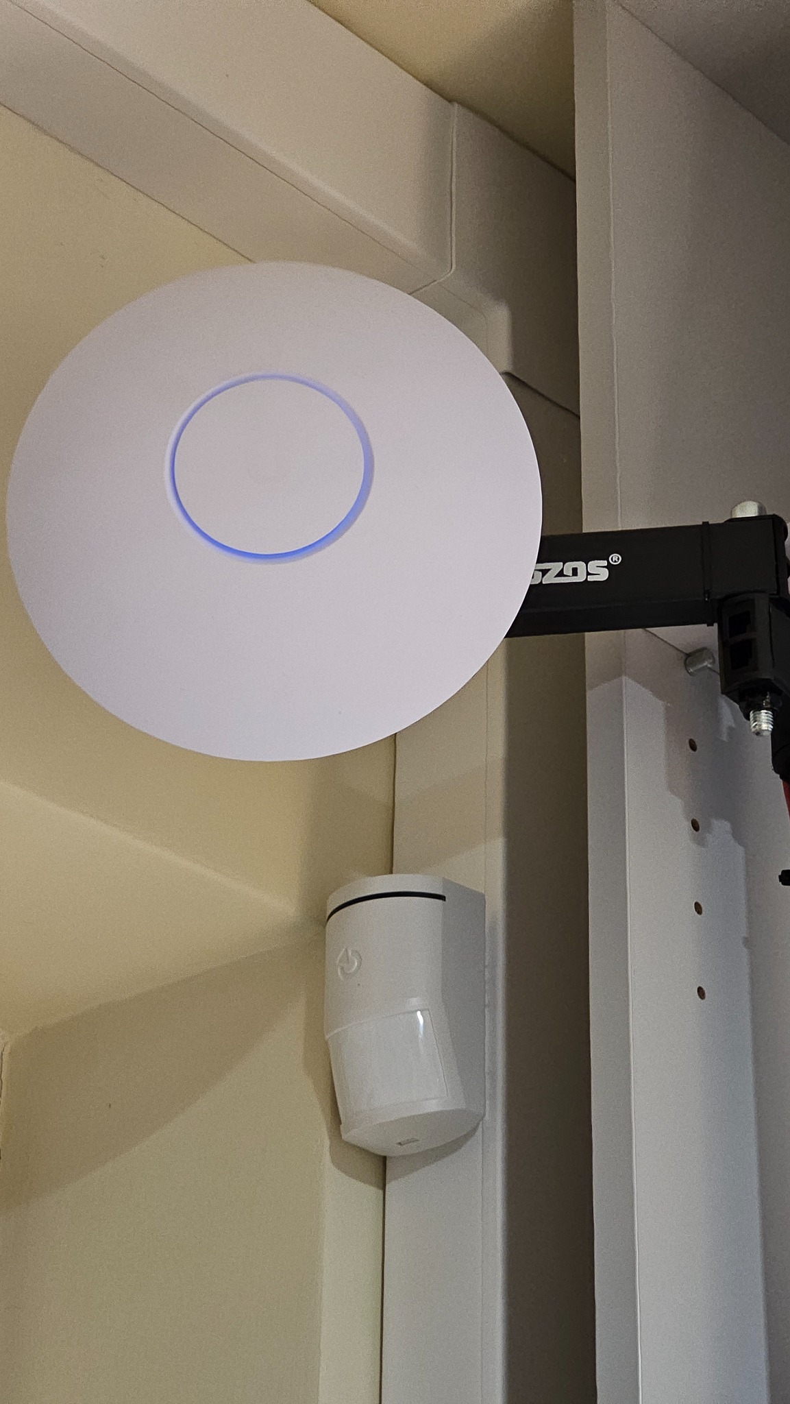 Zamontowany punkt dostępowy Wi-Fi (access point) z niebieskim podświetleniem, obok czujnik ruchu. Widoczne elementy montażowe.