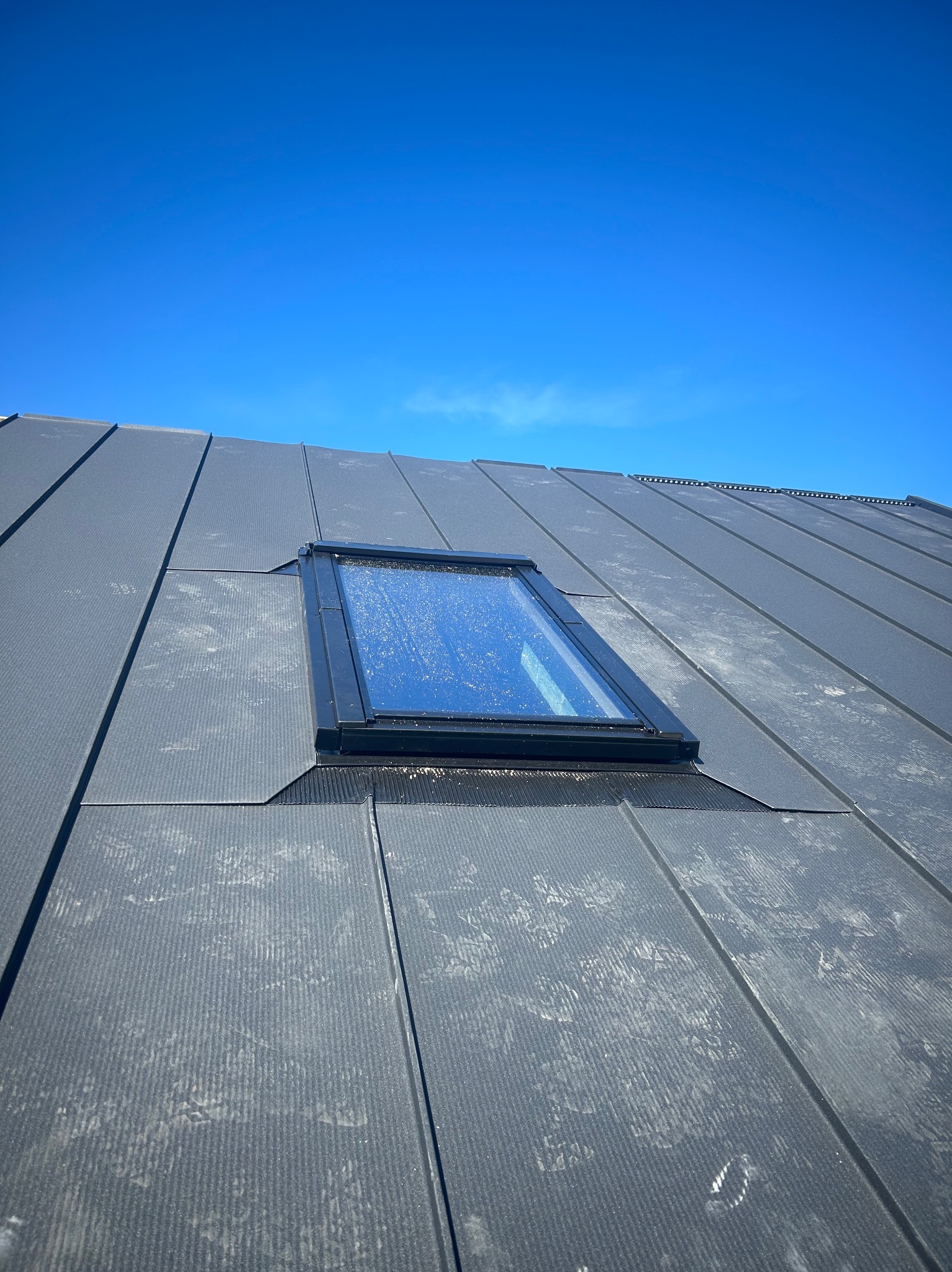 Grafitowy dach z blachy na rąbek stojący, z oknem dachowym, na tle błękitnego nieba. Widoczne ślady po deszczu. Nowoczesne pokrycie dachowe.