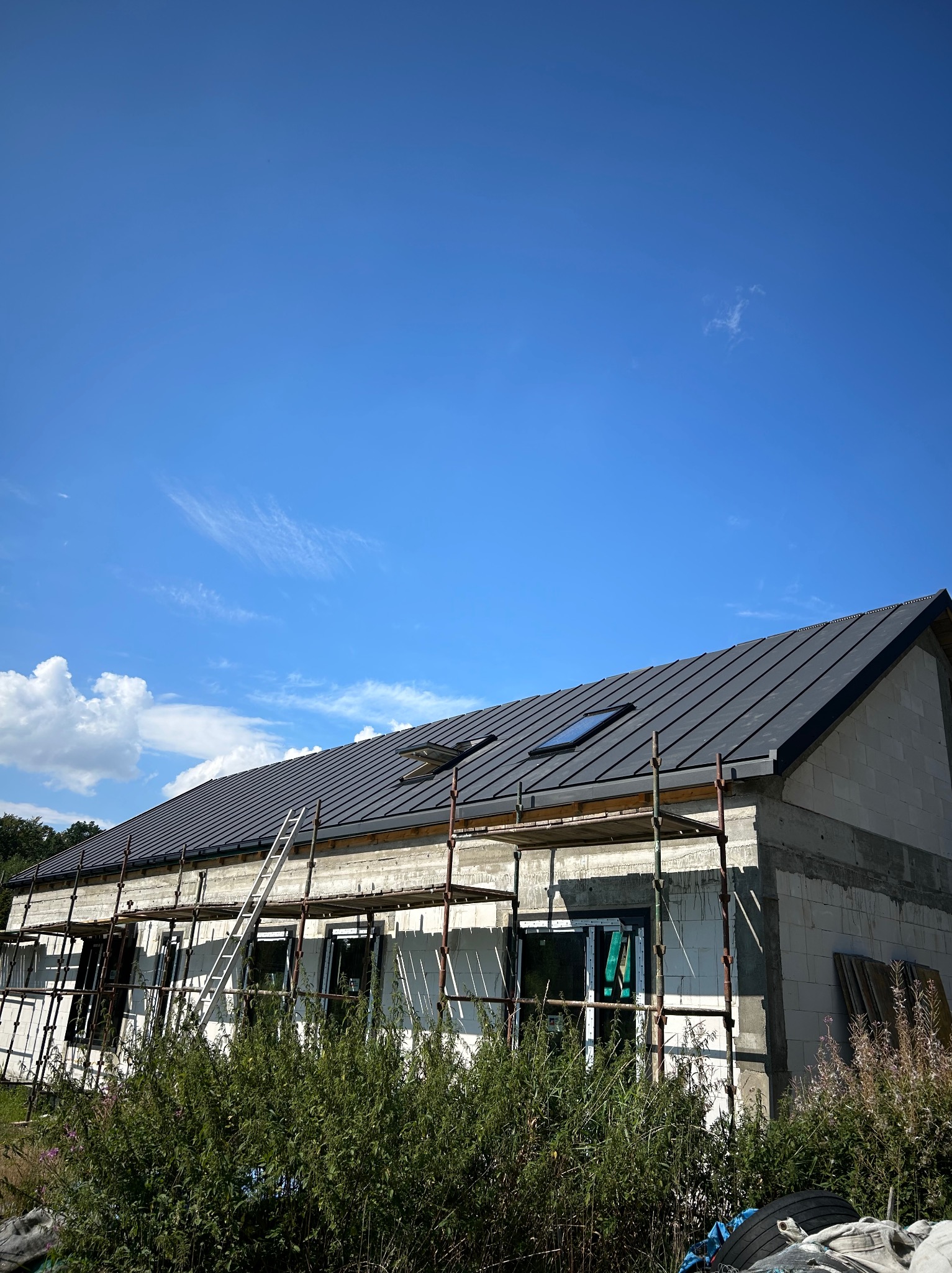 Dom w trakcie budowy z nowym, ciemnym dachem i oknami dachowymi. Widoczne rusztowania i drabina. Błękitne niebo w tle.