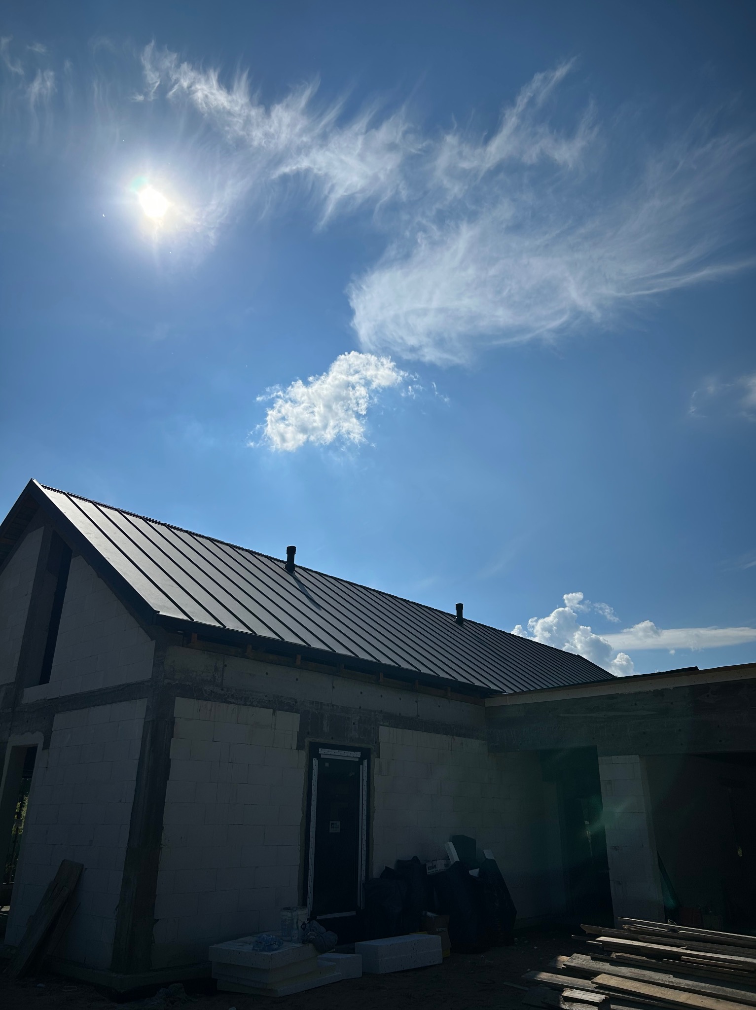 Dom w trakcie budowy z ciemnym, metalowym dachem na tle błękitnego nieba z chmurami. Widoczne materiały budowlane i elementy konstrukcyjne.