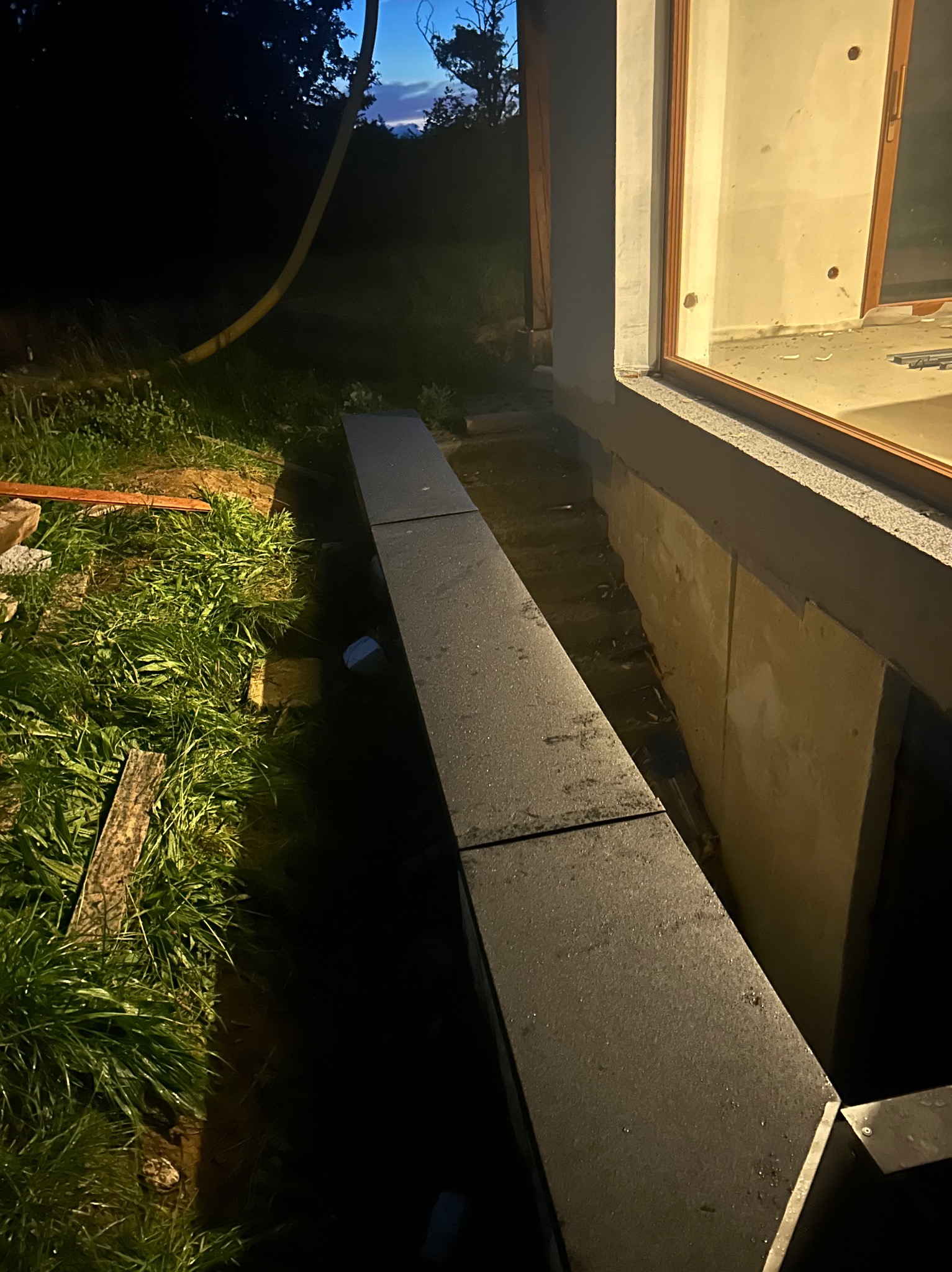 Nocne ujęcie fundamentu budynku z oknem, widoczne czarne płyty izolacyjne i trawa. W tle niebo o zmierzchu i żółty wąż. Prace budowlane w toku.