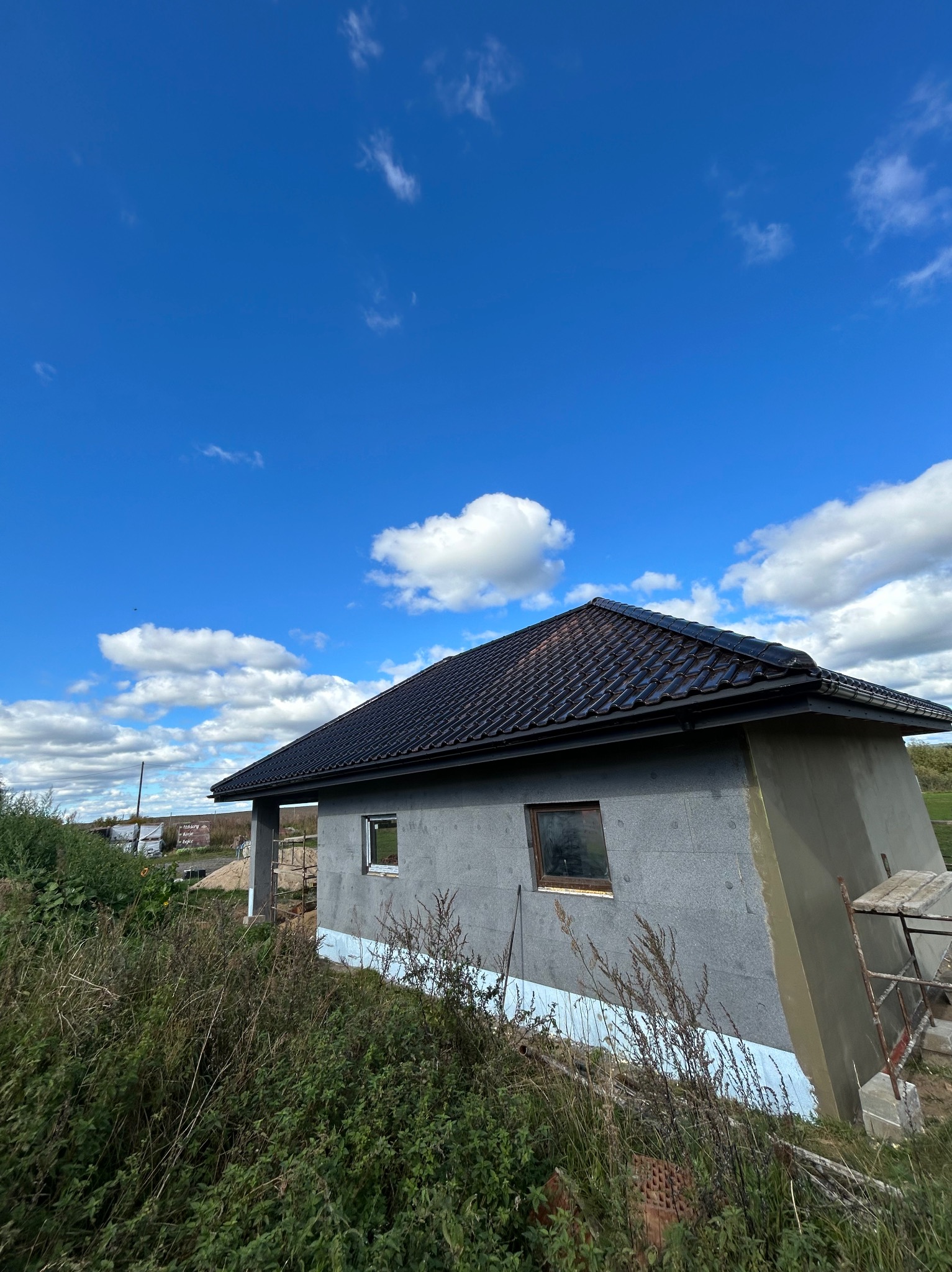 Dom w trakcie budowy z ciemnym dachem na tle błękitnego nieba z chmurami. Widoczne ocieplenie budynku i rusztowanie. Ujęcie z dołu, podkreślające dach.