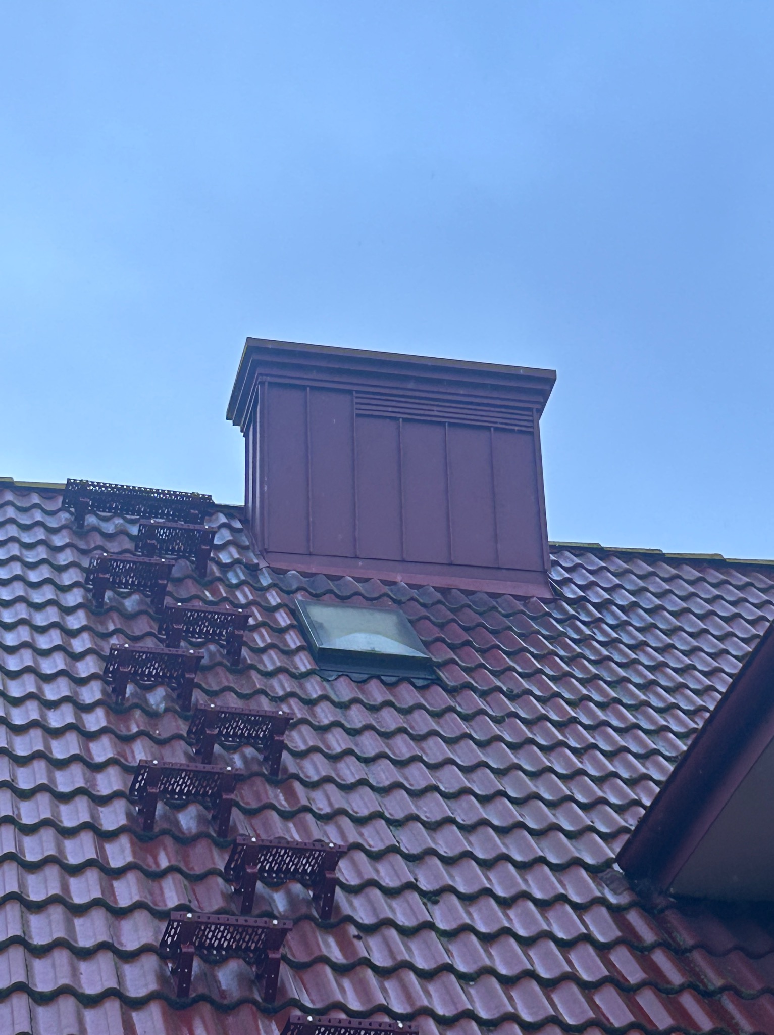 Czerwona dachówka ceramiczna z zamontowanymi stopniami kominiarskimi i oknem dachowym, zwieńczona kominem w kolorze dachówki na tle błękitnego nieba.
