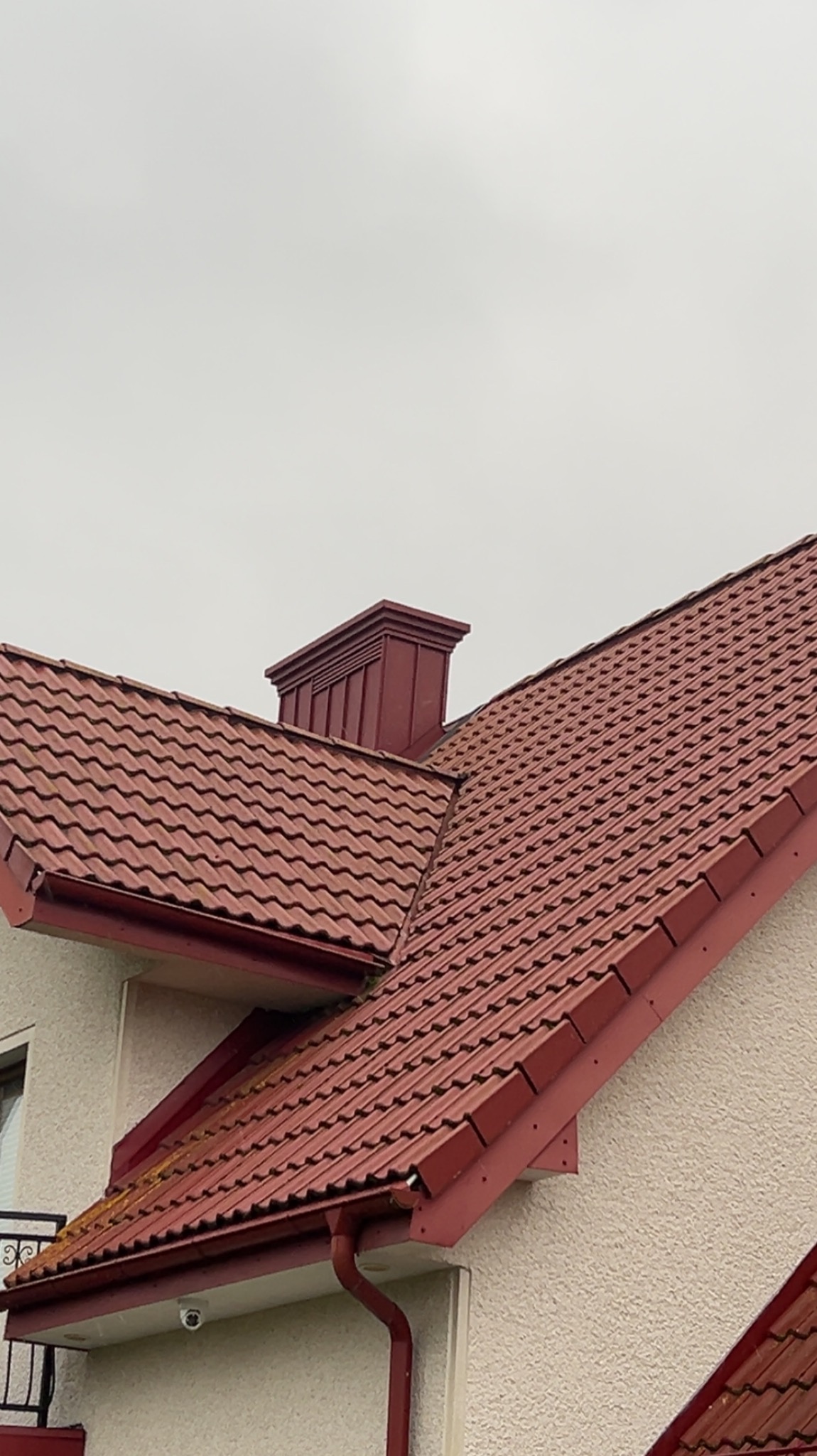 Czerwony dach z dachówki ceramicznej, widoczny komin i rynna. Ujęcie z dołu, fragment elewacji budynku. Widoczna kamera monitoringu.