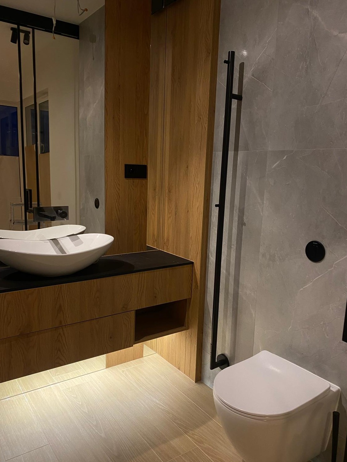 Nowoczesna łazienka z drewnianą szafką pod umywalką, szarą marmurową ścianą i minimalistycznym grzejnikiem drabinkowym. Uwagę przykuwa owalna umywalka nablatowa.