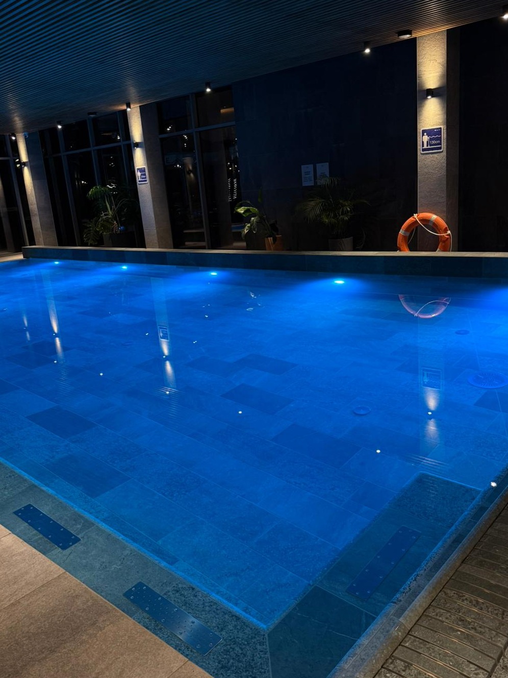 Nowoczesny basen kryty z błękitną wodą, oświetleniem punktowym i roślinami w tle. Pomarańczowe koło ratunkowe na ścianie. Minimalistyczny design.