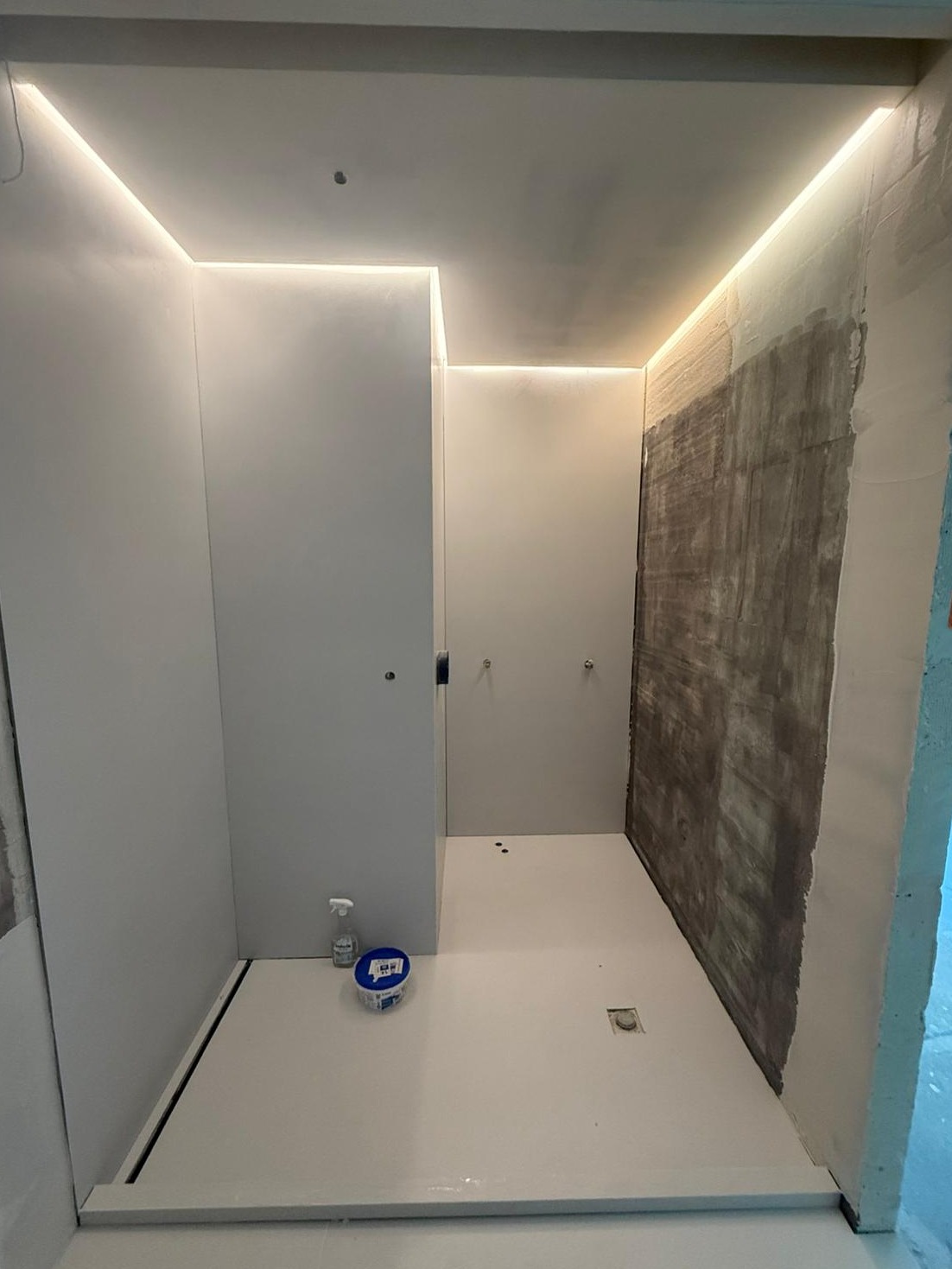 Nowoczesna łazienka w trakcie wykończenia: białe ściany z oświetleniem LED, przygotowana podłoga, ściana w szpachli. Widoczne narzędzia i materiały.