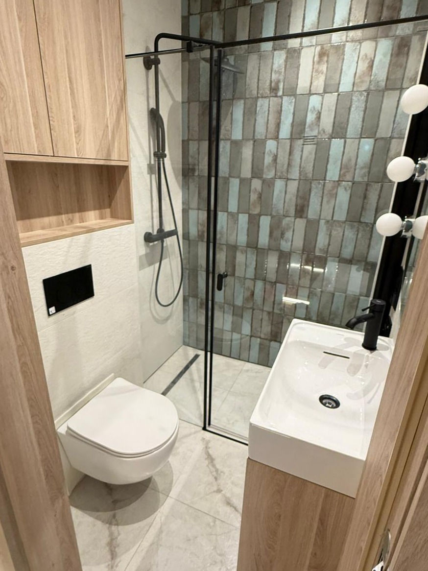 Nowoczesna łazienka z kabiną prysznicową typu walk-in, wiszącą toaletą i umywalką nablatową. Płytki w odcieniach szarości i błękitu. Drewnopodobne szafki.