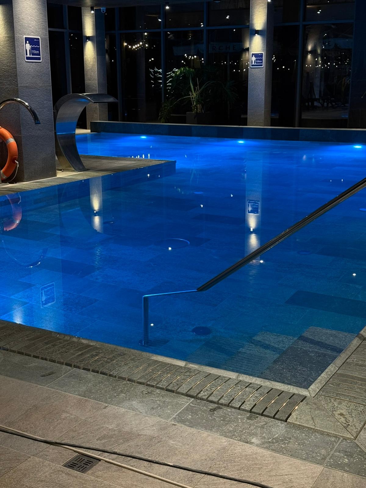 Błękitny basen w spa z kaskadą, oświetleniem punktowym i szarą, kamienną posadzką. W tle okno z widokiem na nocne miasto i rośliny doniczkowe.