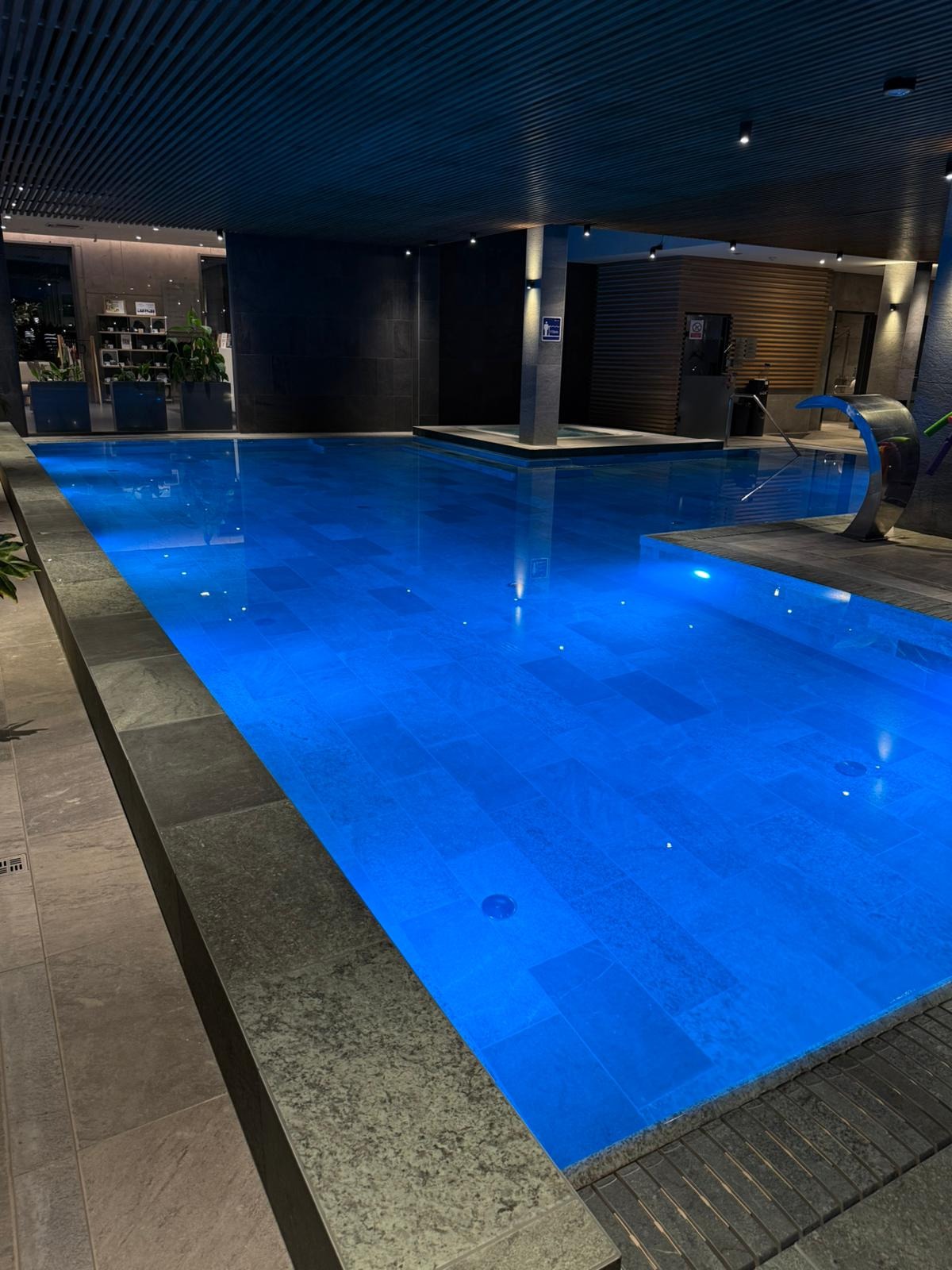 Błękitny basen w nowoczesnym wnętrzu z ciemnymi płytkami i drewnianymi akcentami. Woda oświetlona, tworzy relaksującą atmosferę. Minimalistyczny design.