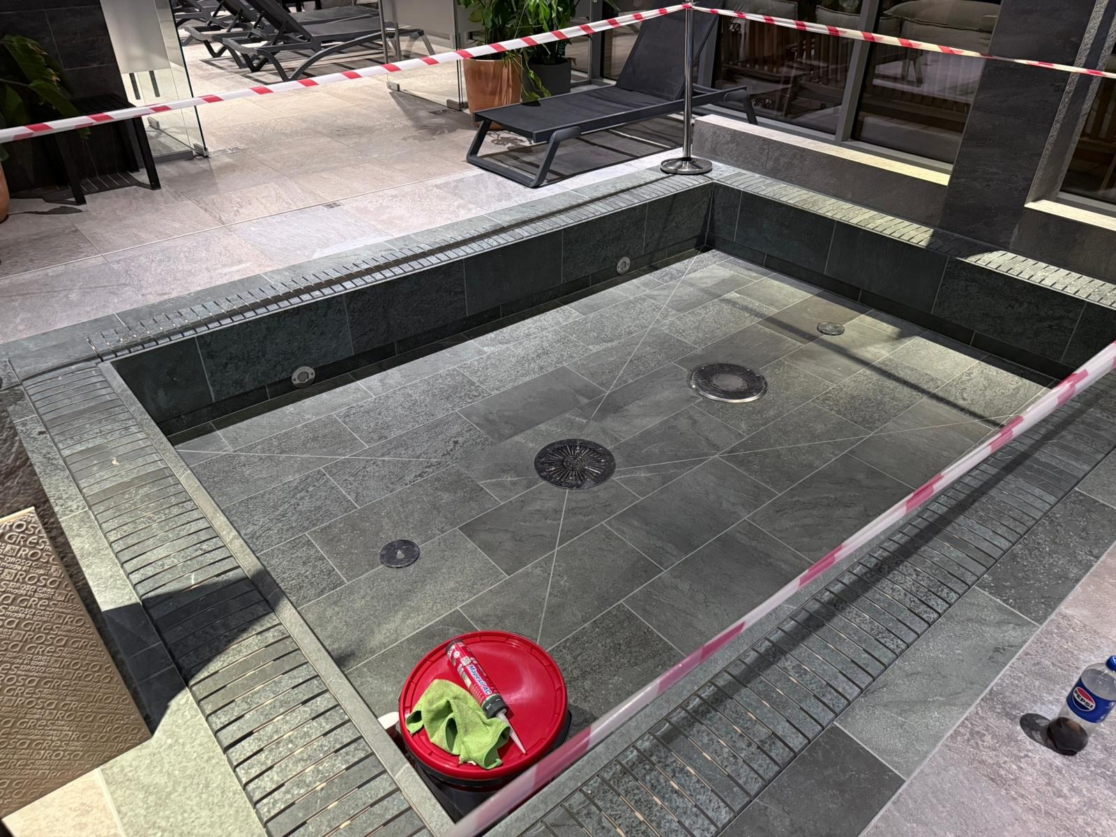 Wykończony brodzik z ciemnoszarą, kwadratową glazurą, zabezpieczony taśmą. Widoczne narzędzia glazurnicze i odpływy. Nowoczesny design.