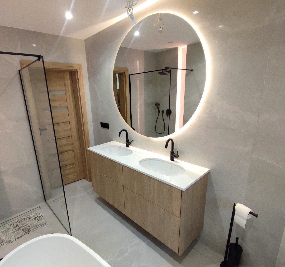 Nowoczesna łazienka z okrągłym lustrem LED, podwójną umywalką na drewnianej szafce i kabiną prysznicową typu walk-in. Minimalistyczny design, szare płytki.
