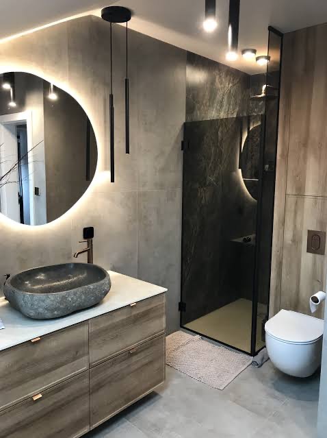 Nowoczesna łazienka z kamienną umywalką, okrągłym lustrem z podświetleniem LED i kabiną prysznicową z ciemnego szkła. Minimalistyczne oświetlenie.