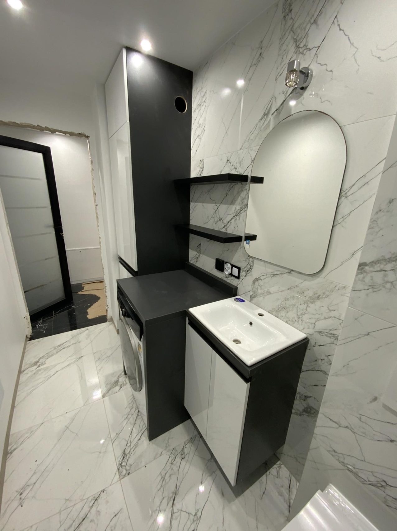 Nowoczesna aranżacja łazienki w Kluszkowcach: białe marmurowe płytki, czarne półki i szafki, umywalka z szafką, pralka w zabudowie i owalne lustro.