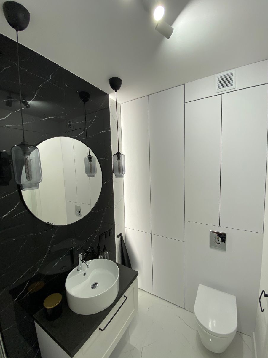 Nowoczesna łazienka z czarnym marmurem, okrągłym lustrem i białymi szafkami. Minimalistyczny design z wiszącą umywalką i toaletą. Oświetlenie punktowe i wiszące lampy.