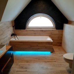 Łazienka na poddaszu z drewnopodobnymi płytkami, podświetlaną wanną i oknem w kształcie półkola. Umywalka z szafką po lewej, toaleta po prawej.