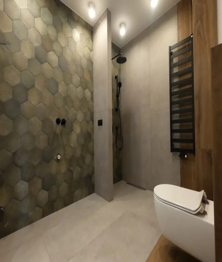 Nowoczesna łazienka z heksagonalnymi płytkami w odcieniach szarości i brązu, czarną armaturą i grzejnikiem drabinkowym. Minimalistyczny design z podtynkową toaletą.