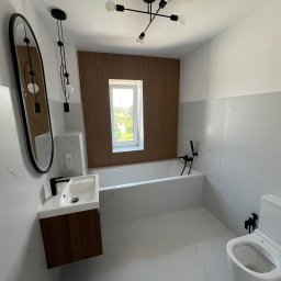 Ra2ns Building - Nowoczesna łazienka z wanną, umywalką z szafką w kolorze drewna i owalnym lustrem. Białe płytki imitujące marmur, czarna armatura i lampa sufitowa.