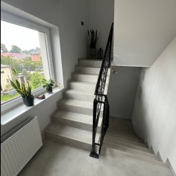 Ra2ns Building - Jasne schody z czarną, ozdobną balustradą w nowoczesnym wnętrzu. Na parapecie okna rośliny doniczkowe. Widok na domy zza okna. Minimalistyczny design.