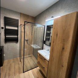 Ra2ns Building - Nowoczesna łazienka z kabiną typu walk-in, drewnianą szafką z umywalką i grzejnikiem drabinkowym. Płytki imitujące drewno na podłodze i szare na ścianach.