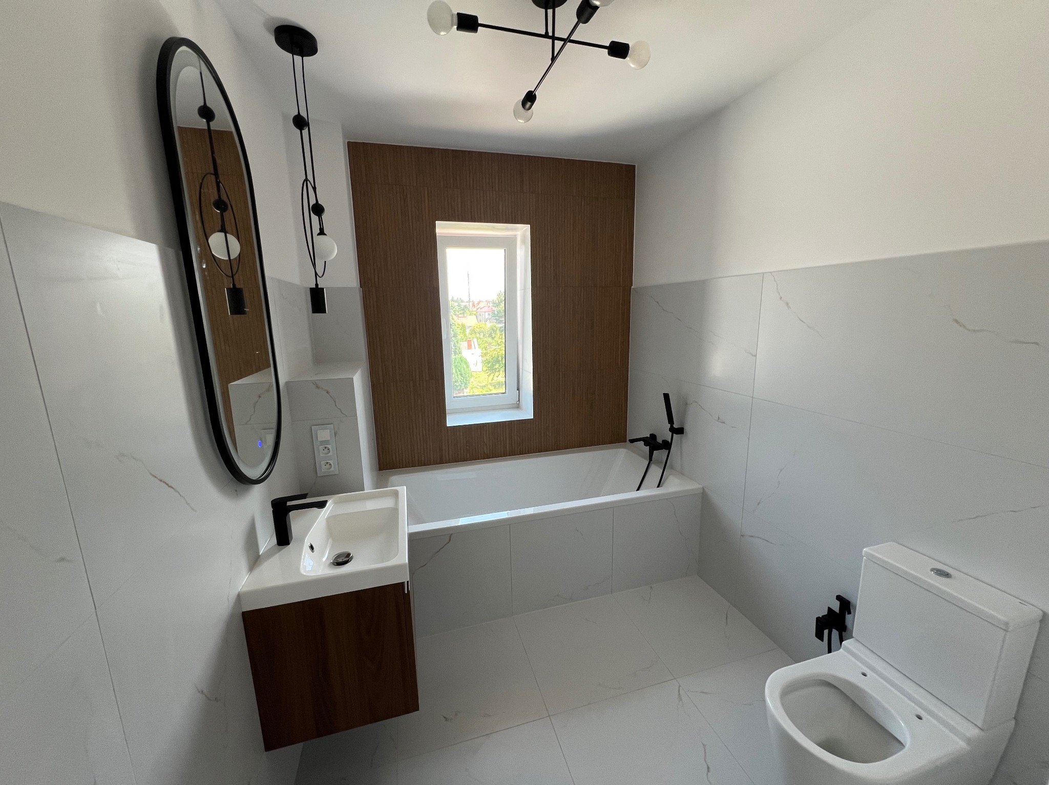 Nowoczesna łazienka z wanną, umywalką z szafką w kolorze drewna i owalnym lustrem. Białe płytki imitujące marmur, czarna armatura i lampa sufitowa.