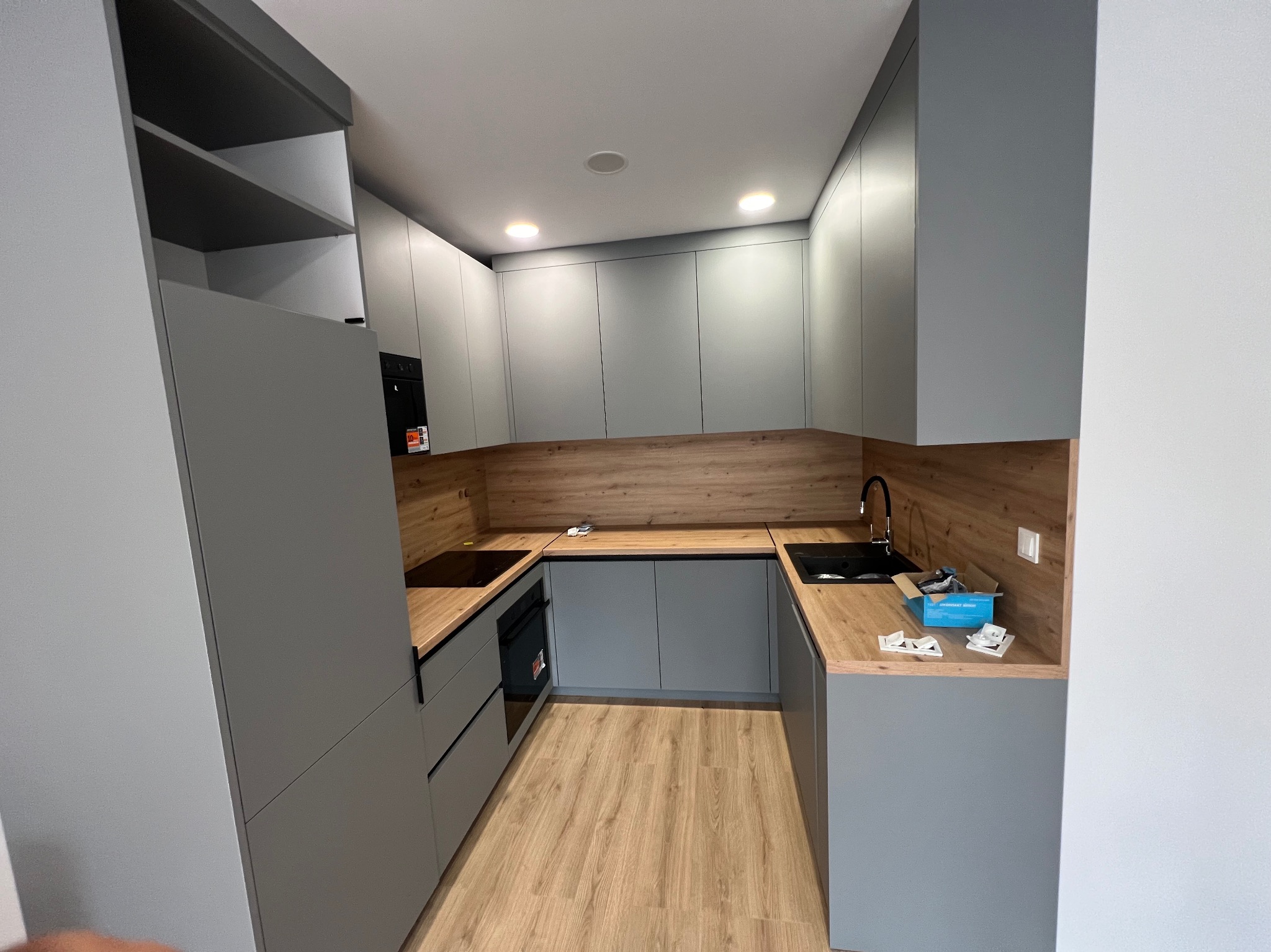 Nowoczesna kuchnia w odcieniach szarości z drewnianym blatem i oświetleniem punktowym. Widoczne szafki, sprzęt AGD i akcesoria montażowe na blacie.