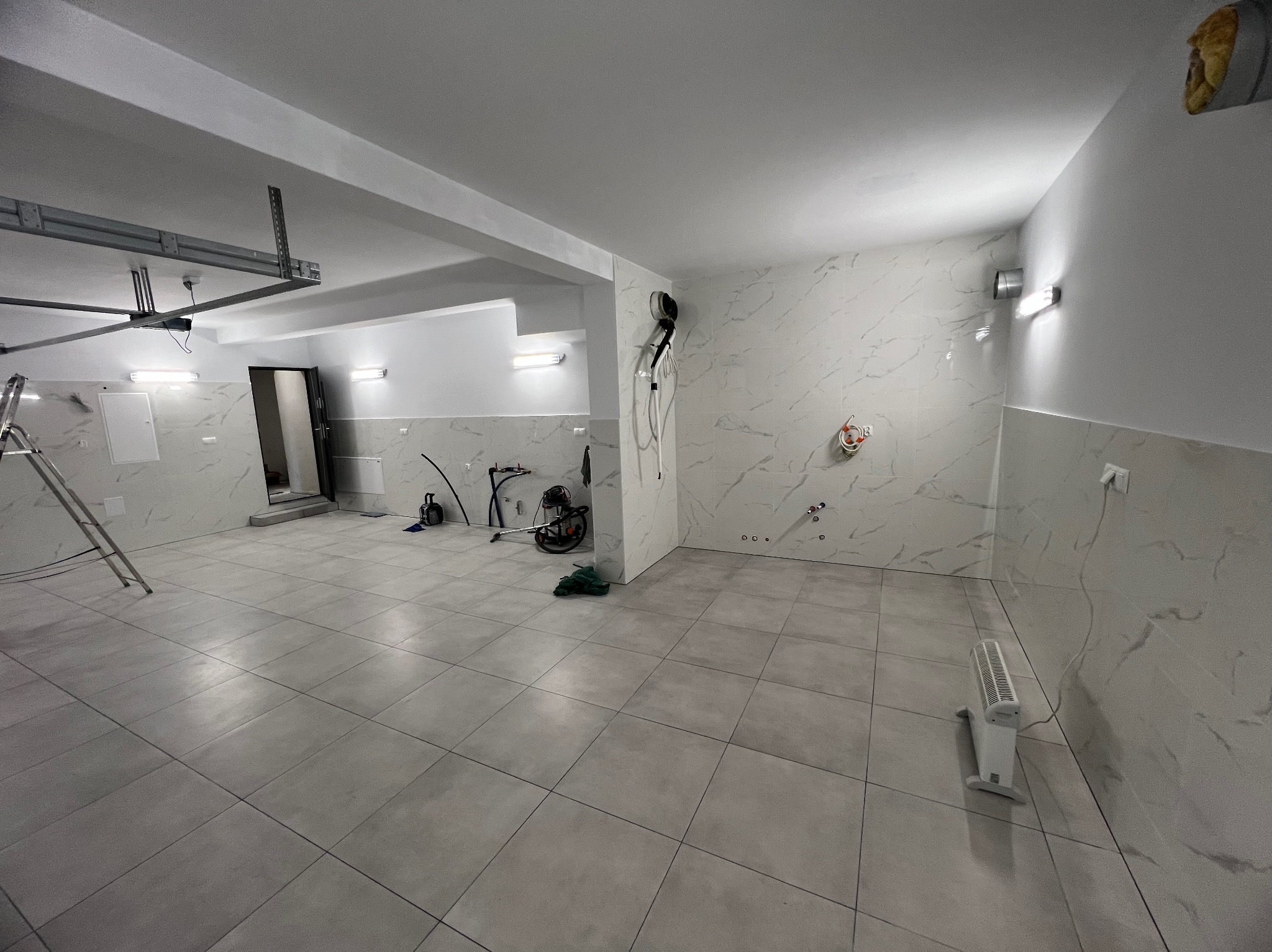 Wykończone wnętrze z marmurowymi płytkami na ścianach i podłodze. Widoczne narzędzia i drabina, sugerujące etap prac wykończeniowych. Oświetlenie punktowe.