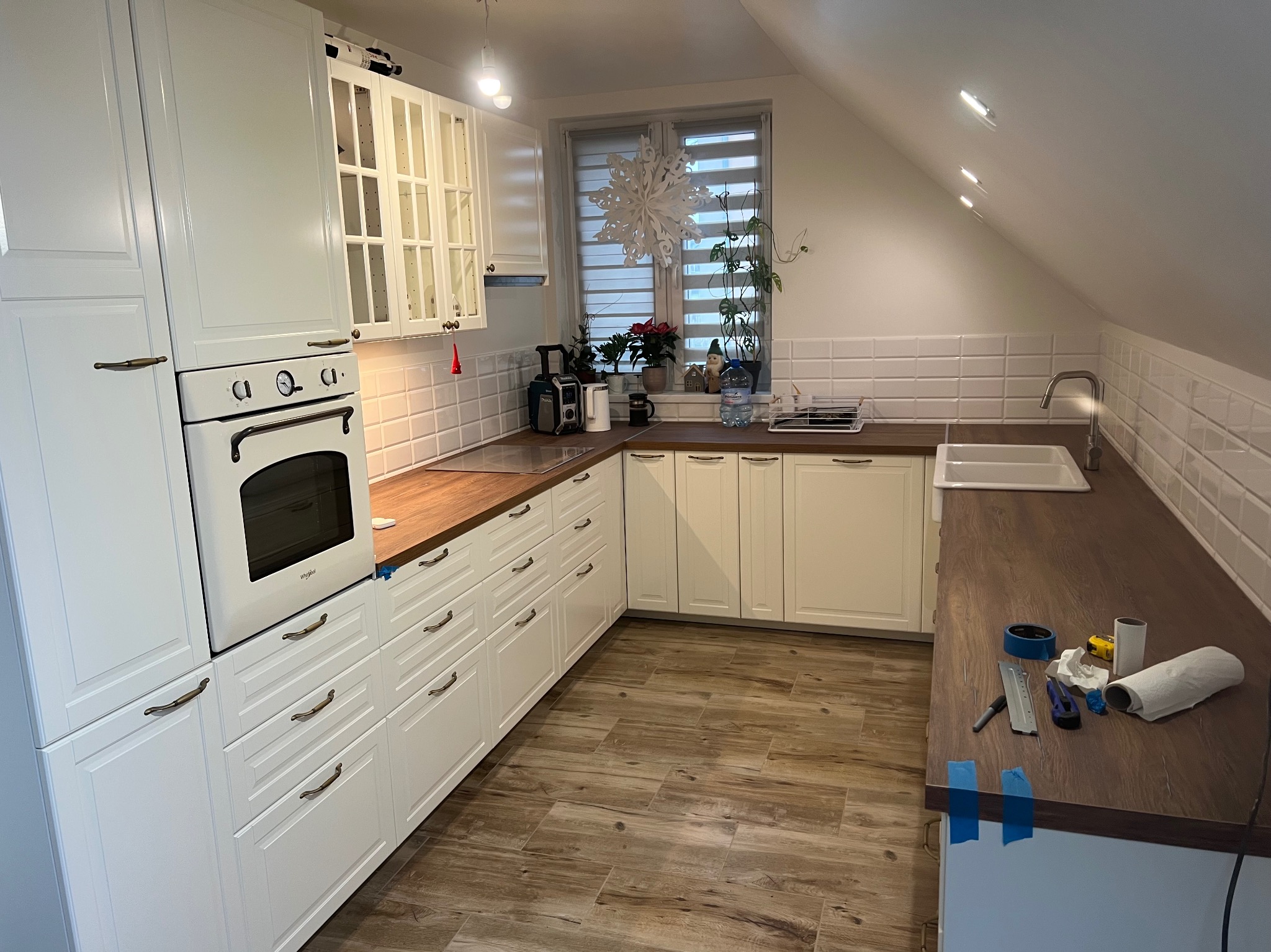 Jasna kuchnia na poddaszu z białymi szafkami i drewnianym blatem. Na blacie widoczne narzędzia, sugerujące prace wykończeniowe. Uwagę przykuwa okno z dekoracją.