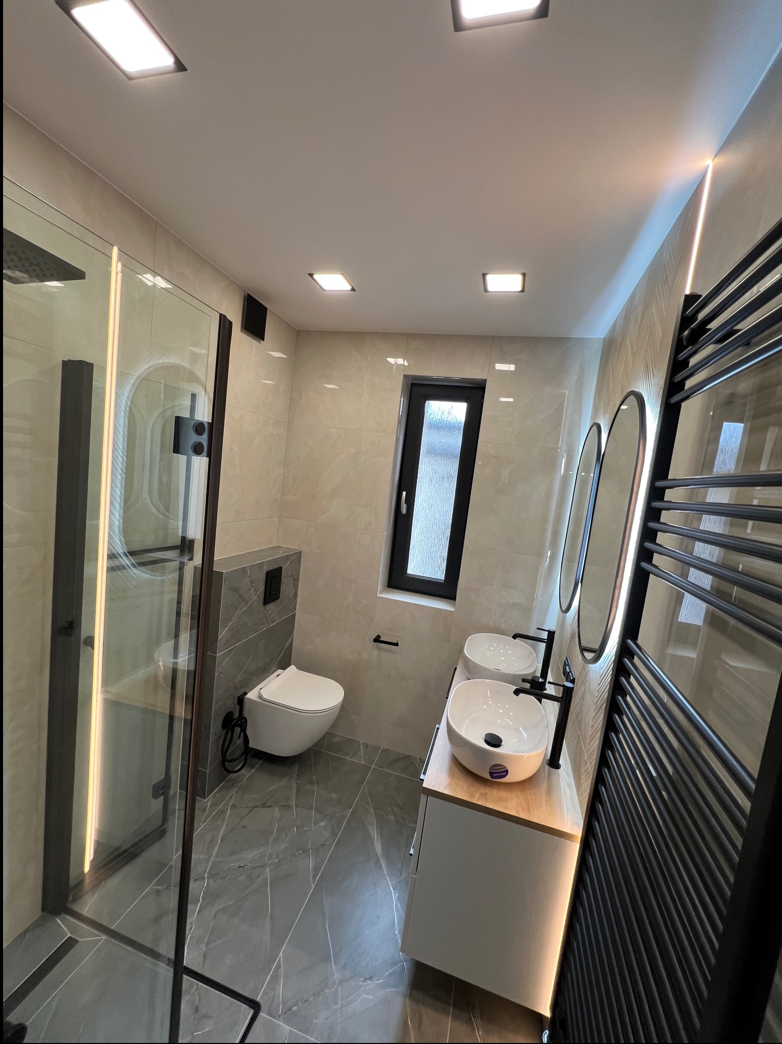 Nowoczesna łazienka z kabiną prysznicową typu walk-in, podwójną umywalką nablatową i grzejnikiem drabinkowym w czarnym kolorze. Uwagę zwraca wąskie okno i oświetlenie LED.