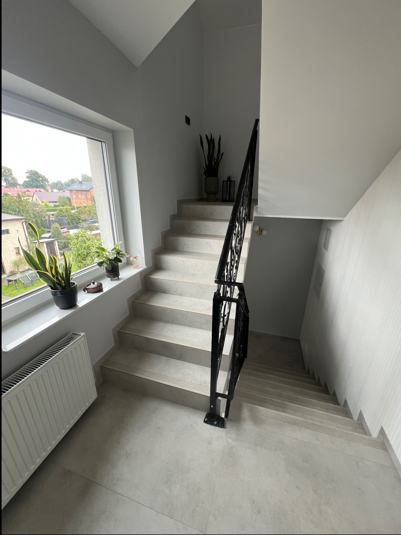 Jasne schody z czarną, ozdobną balustradą w nowoczesnym wnętrzu. Na parapecie okna rośliny doniczkowe. Widok na domy zza okna. Minimalistyczny design.