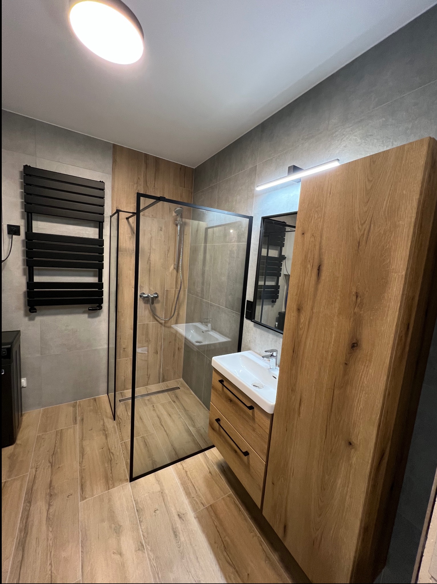 Nowoczesna łazienka z kabiną typu walk-in, drewnianą szafką z umywalką i grzejnikiem drabinkowym. Płytki imitujące drewno na podłodze i szare na ścianach.