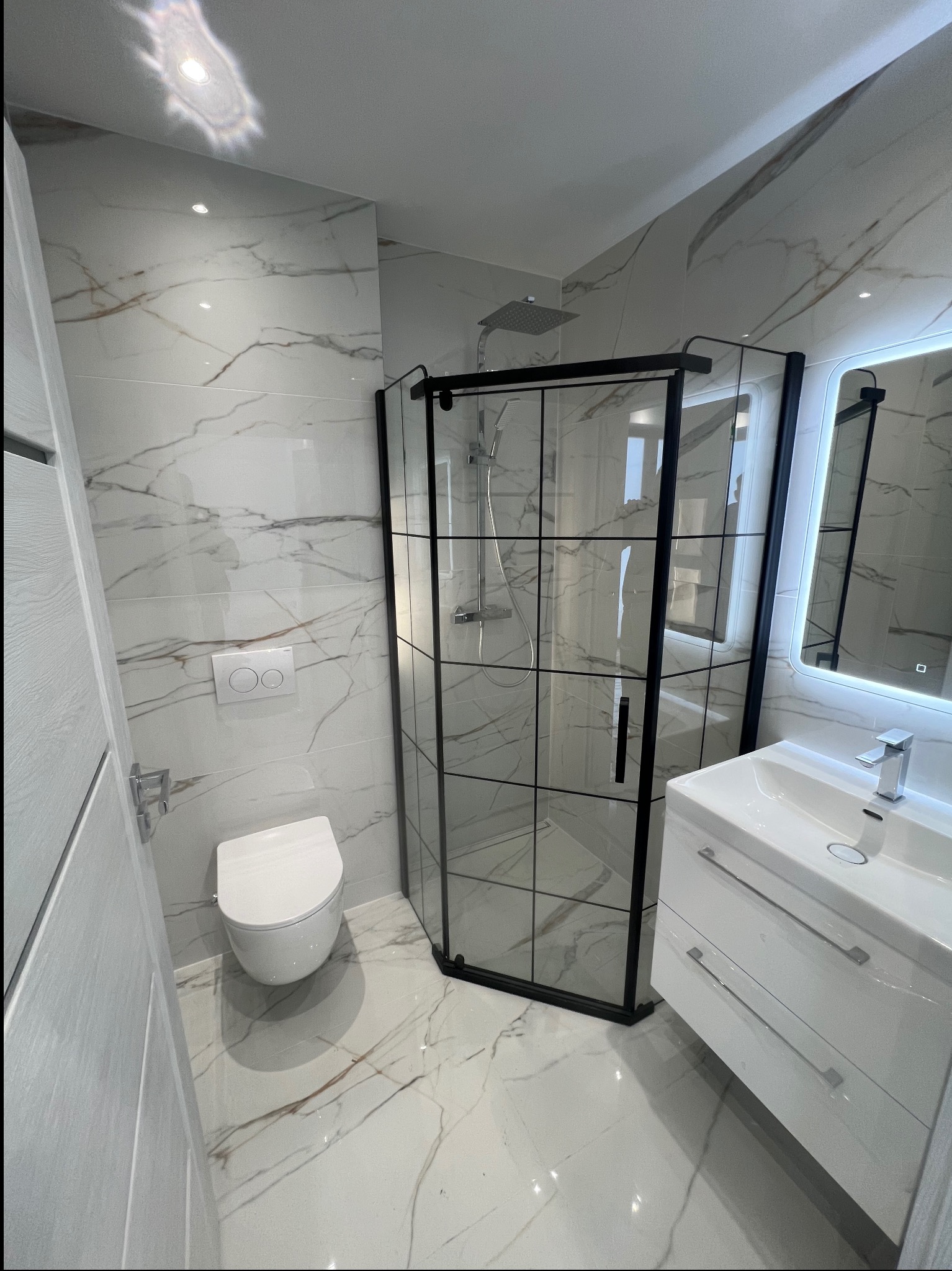 Nowoczesna łazienka z kabiną prysznicową typu walk-in z czarnymi profilami, białą umywalką z szafką i toaletą. Marmurowe płytki na ścianach i podłodze.
