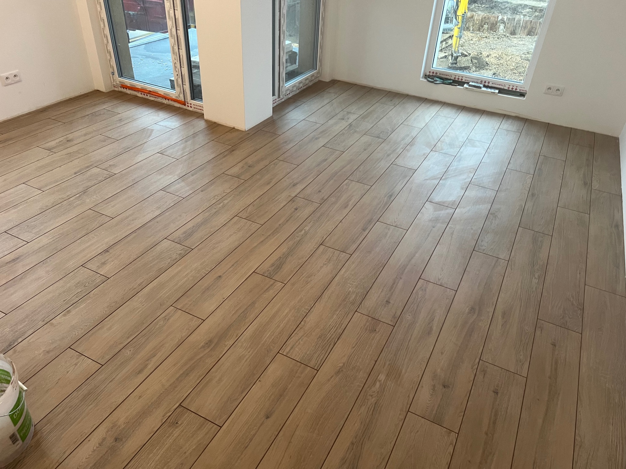 Ułożona podłoga z paneli imitujących drewno w jasnym wnętrzu z oknami. Widoczny fragment wiadra z klejem w lewym dolnym rogu.