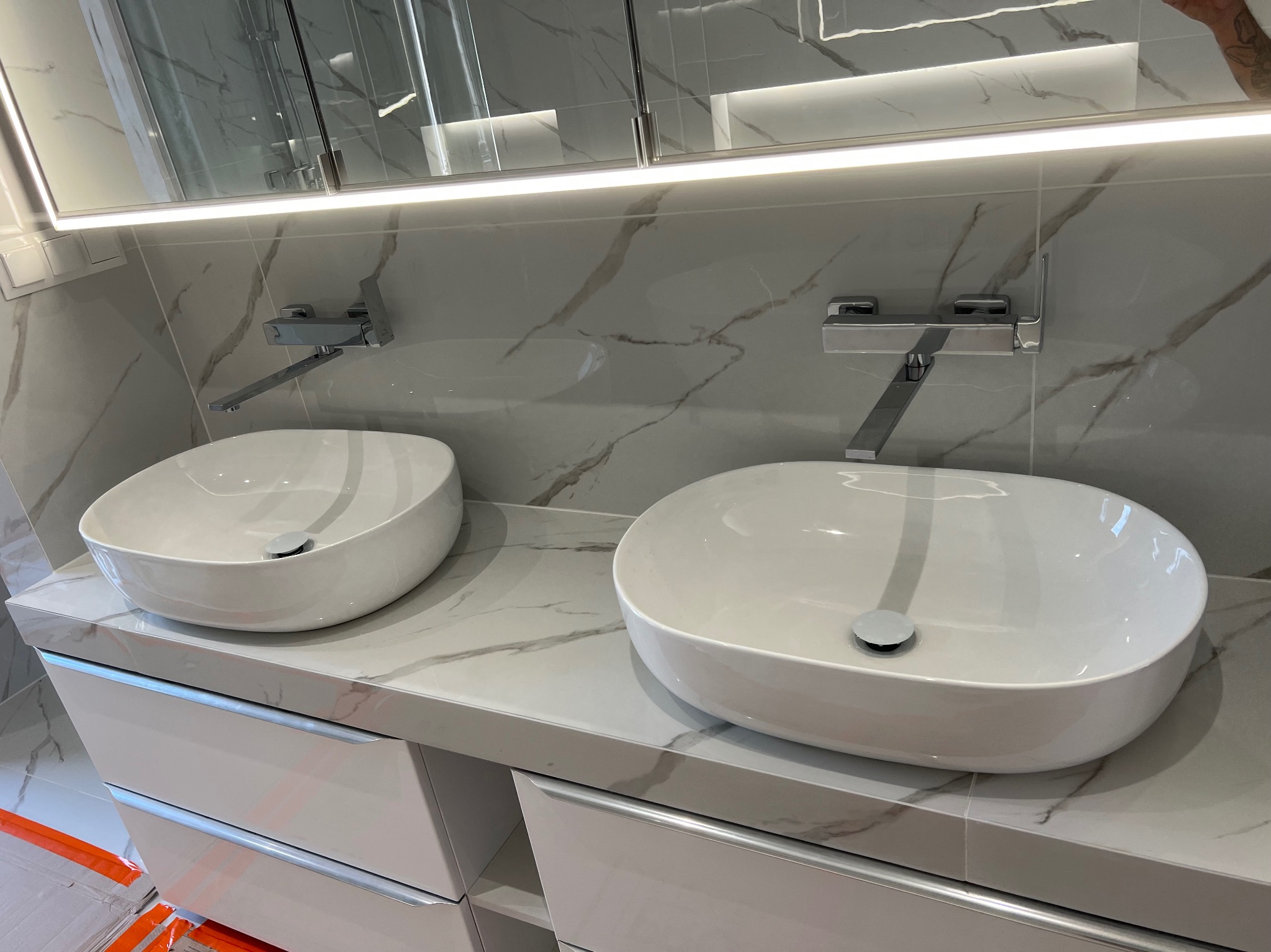 Nowoczesna łazienka z dwoma białymi umywalkami nablatowymi na marmurowym blacie, podświetlane lustro i szare płytki imitujące marmur.