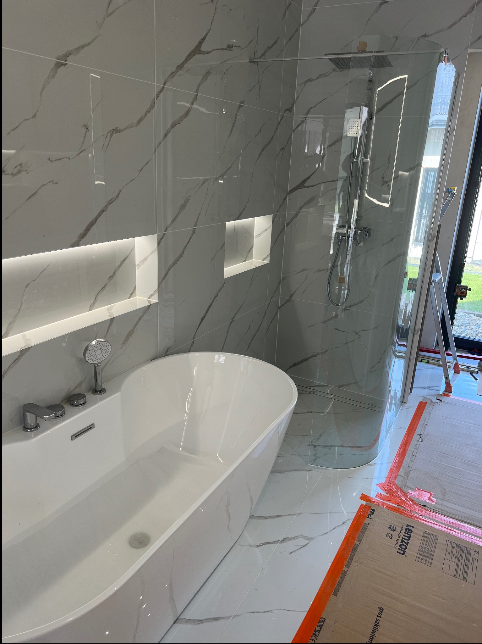Nowoczesna łazienka z wanną wolnostojącą, kabiną prysznicową typu walk-in i płytkami imitującymi marmur z podświetlanymi wnękami. Widoczne narzędzia pracy.