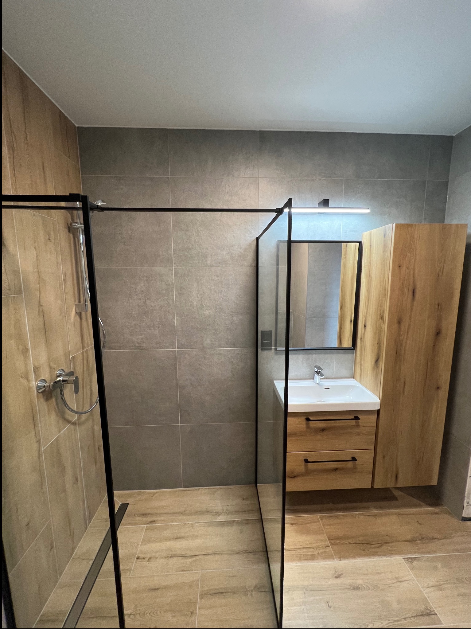 Nowoczesna łazienka z kabiną typu walk-in, drewnianą szafką z umywalką i lustrem. Szare płytki ścienne i podłogowe imitujące drewno. Minimalistyczny design.