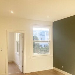 Szymon Różański SELF-MADE - Jasne wnętrze po odświeżeniu: kremowe ściany, okno z widokiem na budynek, akcent w postaci ściany w odcieniu oliwkowej zieleni. Widoczne drzwi wewnętrzne.