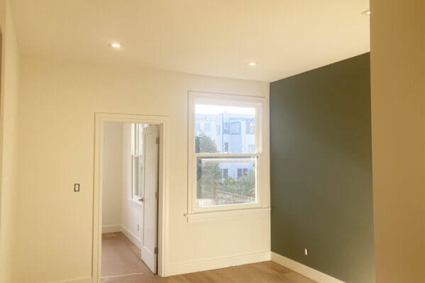 Jasne wnętrze po odświeżeniu: kremowe ściany, okno z widokiem na budynek, akcent w postaci ściany w odcieniu oliwkowej zieleni. Widoczne drzwi wewnętrzne.